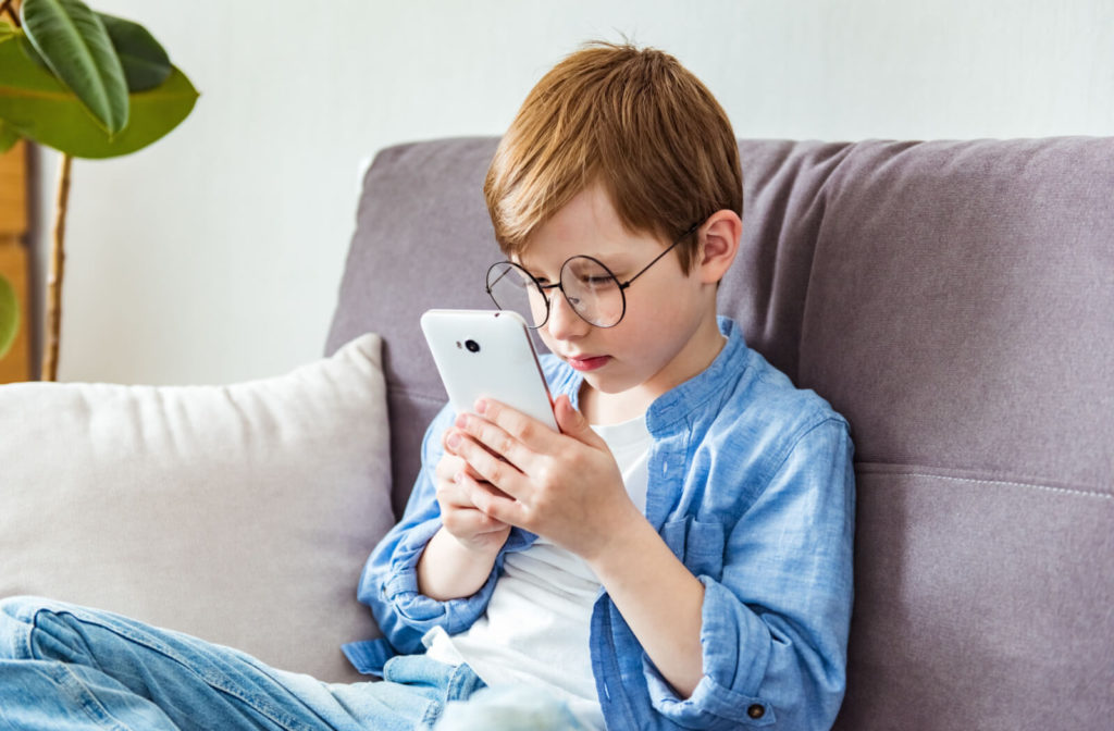 A child with glasses sitting on the couch holds his smartphone very close to his eyes.