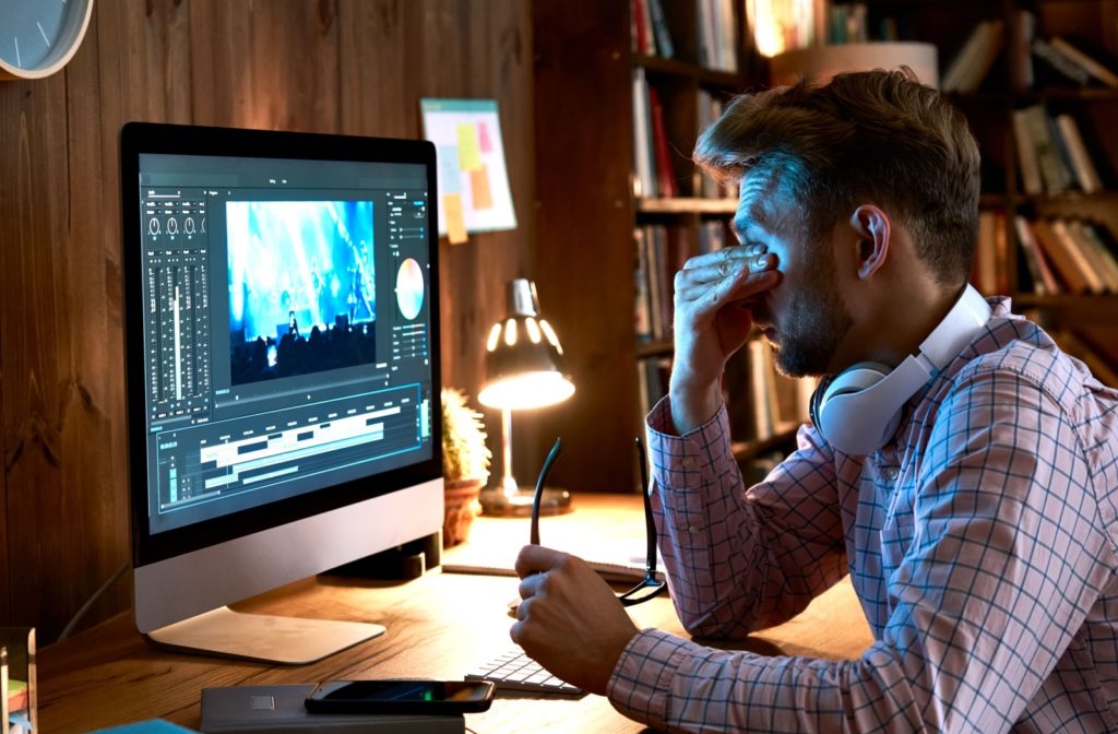 Man rubbing his eyes while working on a computer, holding blue light glasses.