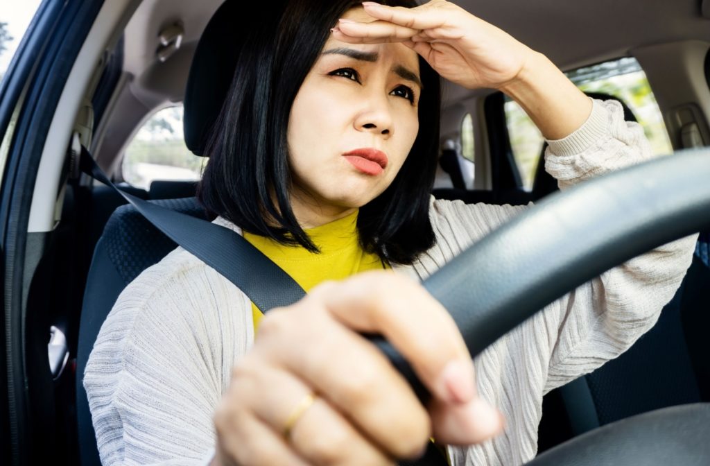 A woman driving in her car lifts her left hand to shade her eyes as she squints to see the road due to blurry vision.