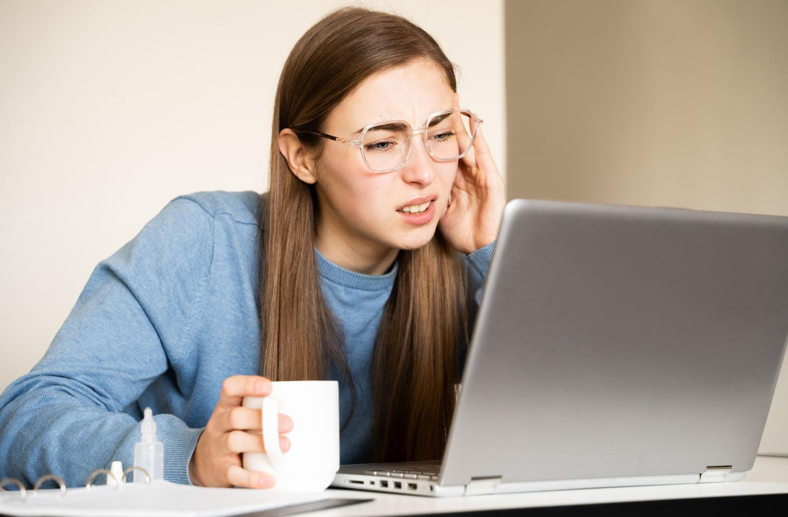 A person squinting and holding the side of their head while looking at a laptop screen, experiencing double vision due to eye strain.